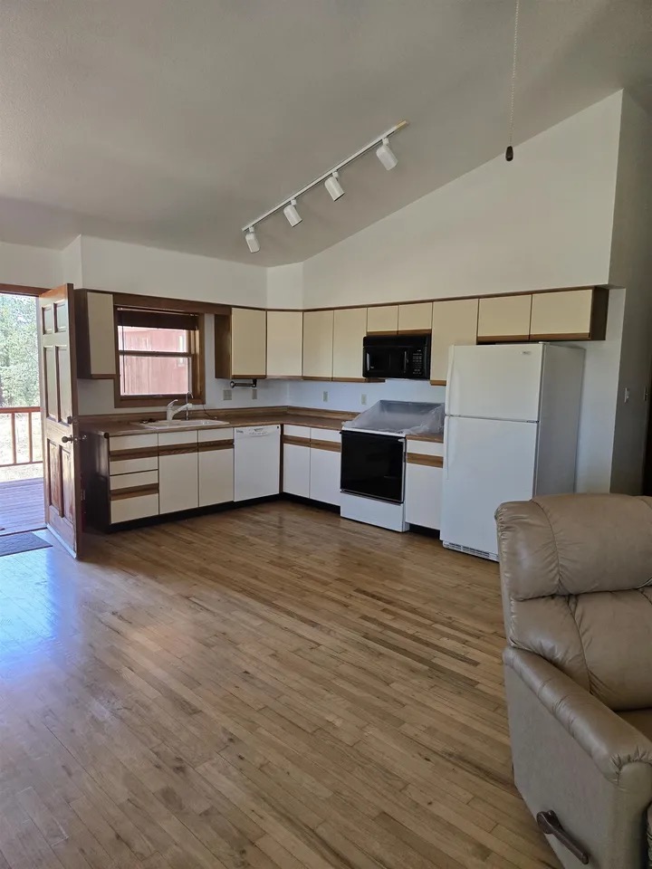 Open kitchen and great room with a vaulted ceiling, track lighting, white cabinets, light wood floors, and a door opening onto the deck