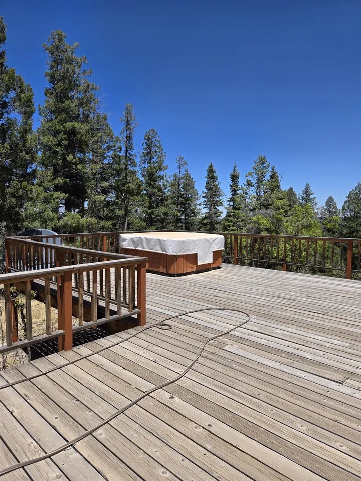 The wide wood deck running the length of the home, a covered hot tub at the far railing and tall pines beyond under a clear blue sky